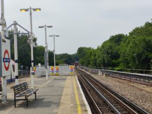 Looking along the length of another station platform with trees the other side of the track, on the right this time.  A tube train is approaching in the distance.